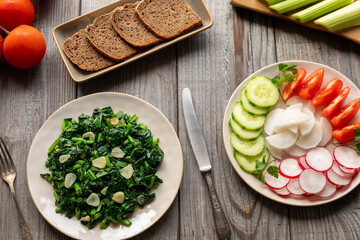 A woman mixes rocket leaves and quinoa salad. Main ingredients in bowls: quinoa, tomato, radish, and celery and olive oil dressing in a glass bottle. Green houseplant behind.
