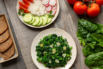 Colorful vegan meal. Stir-fried spinach leaves with garlic, plate with fresh-cut vegetables, and plate with slices of homemade rye bread. Whole tomatoes and fresh spinach leaves on the side.
