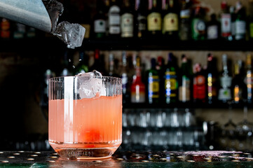 Dynamic ice cubes splashing into a pink cocktail at a well-stocked bar. The photo captures the moment of preparation, with the glass placed on the bar counter amidst an array of liquor bottles.