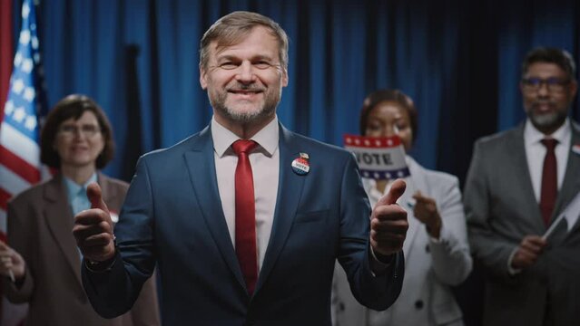 Medium portrait shot of cheerful Caucasian male candidate for presidency, Senate or Congress, showing thumbs up, smiling, and multiethnic team waving vote flags and cheering