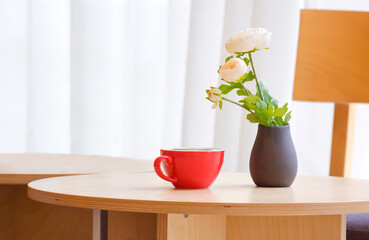 Hot latte art coffee in red mug with artificial flowers in clay vase on round wooden desk with blurred background of white curtain inside of modern coffee shop, close up with copy space