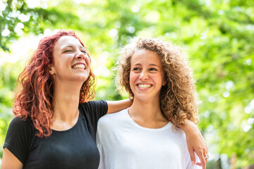 Two happy women with curly hair laughing and hugging in a sunny park - 754221021