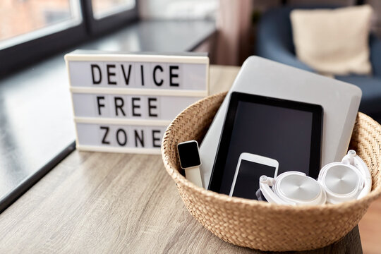 digital detox and technology concept - close up of device free zone words on light box and different gadgets in wicker basket on table at home
