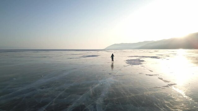 A woman in a blue down jacket skates on the blue transparent ice of the endless winter lake Baikal at sunset