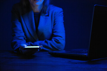 Business woman holding a smartphone and working on a laptop in the dark and illuminated by the...
