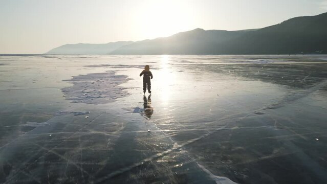 A child in a down jacket is clumsily skating on the blue transparent ice of the endless winter Lake Baikal at sunset