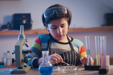 A modern young chemist in a home research laboratory wearing safety glasses is intently conducting...
