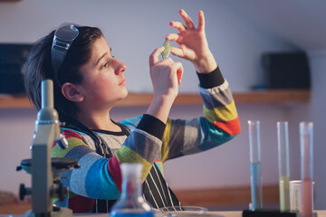Cute young botanist in a home research laboratory wearing safety glasses is intently studying a plant leaf on a microscope slide. Advanced school science project in botany - obrazy, fototapety, plakaty
