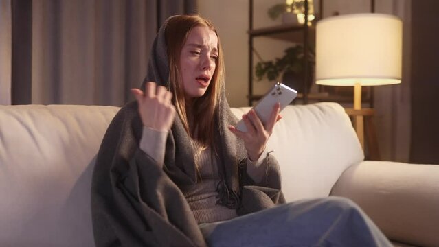 Portrait of crying young blond woman with smartphone sitting on the bed under the blanket and reading bad news email break up message from ex-boyfriend or personal problems at home at late night