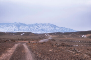 Fototapeta na wymiar Dirt road through mountain steppe to snowy mountains in early spring in Kazakhstan
