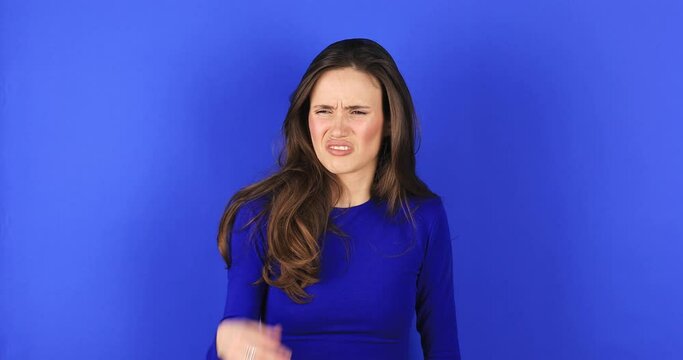 Disgusting odor. Discontent brunette woman frowning face, smells something awful, feeling aversion, blue studio background. Girl face with aversion dislikes something unpleasant. Woman wear blue top.