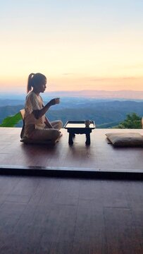 A woman is relaxing on a wooden floor, gazing out a window at the sunset over a tranquil lake. The sky is painted with vibrant colors, creating a stunning landscape art piece in Northern Thailand