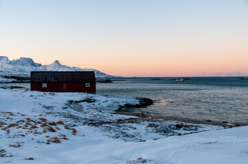 A traditional norwegian red barn enclosed by rugged mountains and snow-covered beaches in the arctic. Shown during the golden hour of a brief period of daylight near the town of Bodo.