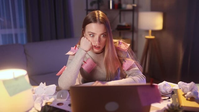 Nervous stressed blond young woman freelancer or student with stickers typing quickly on computer and looking at papers asking to hurry up and warning about deadline at home workplace late at night