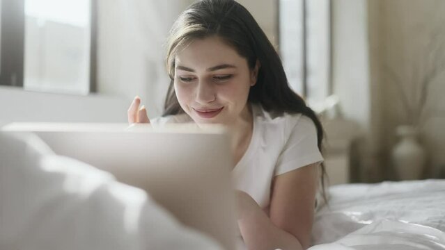 Portrait of positive young woman thinks questioningly imagine plan in mind has doubts create idea while finding solution idea with happy inspired face laying on bed with laptop computer at home