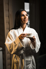 Young woman in a white bathrobe drinks espresso coffee in a wooden hotel room. Hotel holiday...