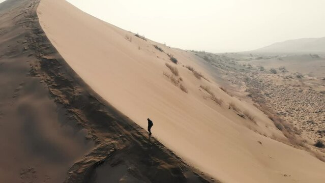 A man climbing up the ridge of a sand dune to the top. Arid desert terrain. Cold season. Shooting from a drone. Singing Barkhan. Altyn-Emel National Park