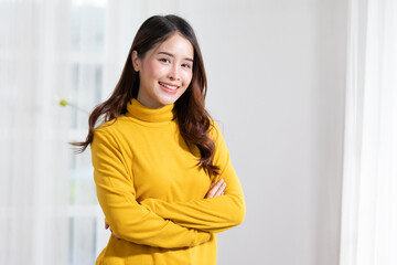 Radiant young woman smiling in a fashionable yellow turtleneck, arms crossed in a bright room