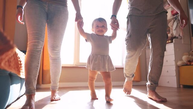baby with parents first steps. happy family kid a dream concept. baby daughter takes first steps holds family hands silhouette against window indoors. cute baby toddler learning walk lifestyle