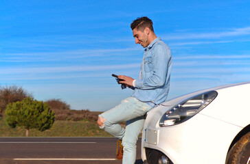 Curly haired Caucasian man leaning on his car smiling, using mobile phone, with the road beside, for copy space, side view. Lifestyle concept