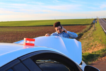 Caucasian man calling on the phone looking at the engine with the emergency light V16, daytime. Concept of problem on road