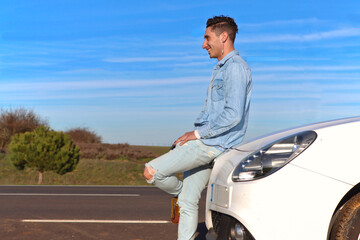 Curly haired Caucasian man leaning on his car smiling, looking at the horizon, with the road in the background, for copy space, side view. Lifestyle concept