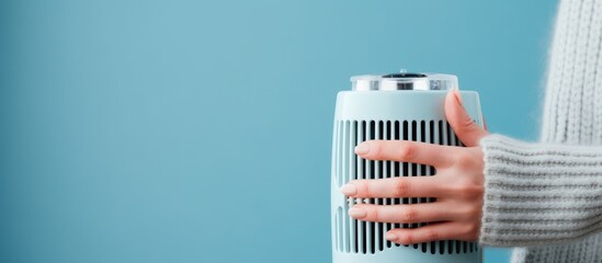 A woman is holding a blue air conditioner in her hands against a blue background. The portable domestic appliance is used for cooling during hot weather. It is a front view image.