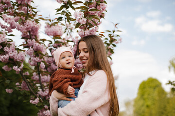 Obraz premium A teenage girl with long hair in a pink jacket is holding a small child near a blooming cherry blossom. The older sister is holding his little sister and smiling near the cherry blossom. 
