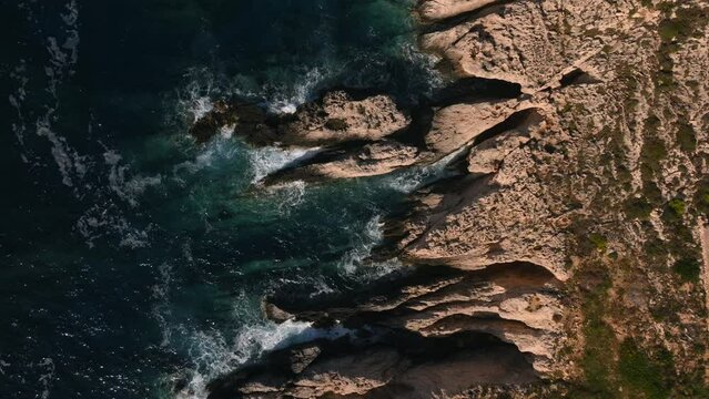 Aerial top down view of waves crashing into rocky sea cliffs during sunset golden hour