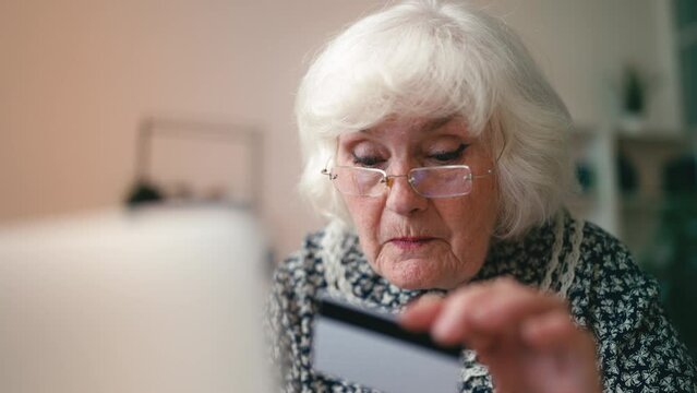 Funny granny in glasses shopping online, entering credit card number on laptop