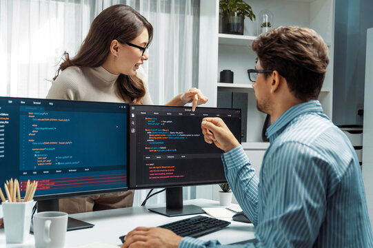 Woman colleague standing opposite to man programmer working on desk to discuss coding website software system project on two pc screen, comparing with typing full stack data prompt design. Postulate.