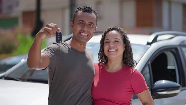 Young hispanic couple hugging each other holding key of new car at street
