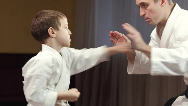 A little athlete with a white belt performs punches on the palms of the trainer