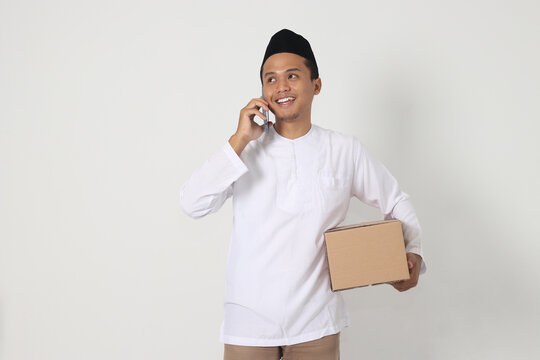 Portrait of happy Asian muslim man in koko shirt with peci carrying cardboard box while having having conversation on mobile phone. Going home for Eid Mubarak. Isolated image on white background