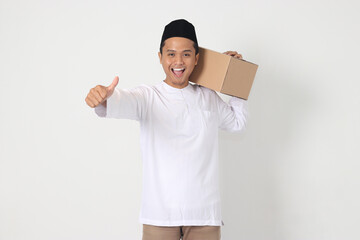 Portrait of excited Asian muslim man in koko shirt with peci carrying cardboard box while pointing...