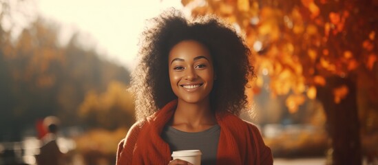 A cheerful African woman in casual attire smiling, holding a cup of coffee against an autumn nature background on a sunny day. She enjoys her coffee break outdoors with a beautiful smile.
