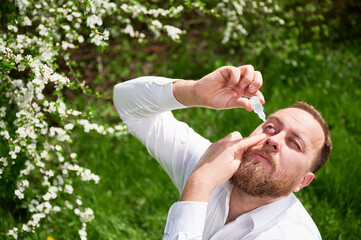 Man allergic using medical eyes drops, suffering from seasonal allergy at spring in blossoming...
