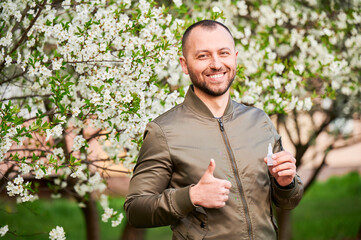 Man allergic using medical eyes drops, suffering from seasonal allergy at spring in blossoming garden. Portrait of smiling man showing thumbs up in front of blooming tree. Spring allergy concept.