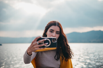 A young woman with dark hair captures a selfie on her smartphone against a scenic lake backdrop