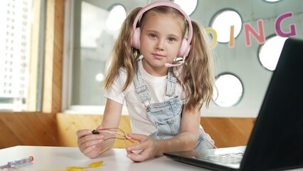 Girl wearing headphone while study equipment and looking at camera. Caucasian child doing science experiment with laptop, screwdriver and wires placed near on table. Smart online classroom. Erudition.