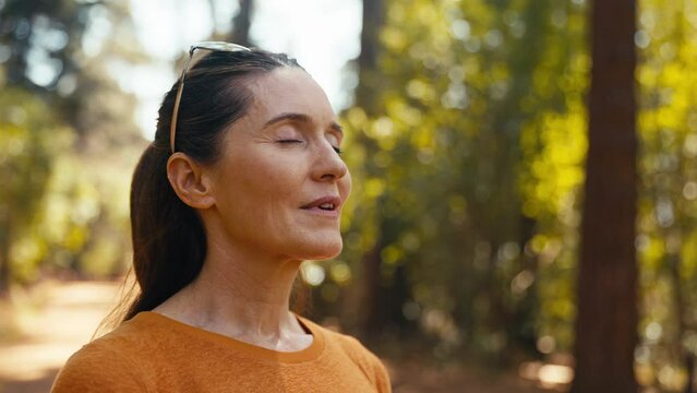 Peaceful senior retired woman relaxing standing amongst nature in forest surrounded by trees with closed eyes breathing deeply - shot in slow motion