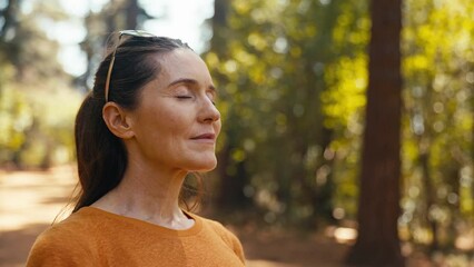 Peaceful senior retired woman relaxing standing amongst nature in forest surrounded by trees with closed eyes breathing deeply - shot in slow motion