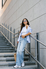 Young woman in a blue shirt and jeans stands on a set of stairs. She is wearing sunglasses and a white shirt