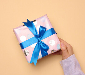 A woman's hand holds a gift box wrapped in a blue silk ribbon on a beige background