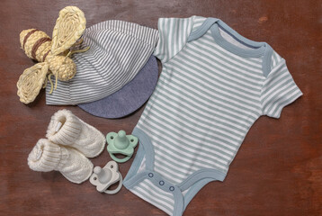Flat lay of blue striped baby boy onesie and cap with white booties and pacifiers on brown wood background with copy space for gender reveal or announcement