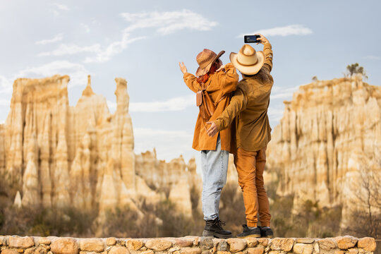 Two people are taking a picture of a mountain range. The man is wearing a hat and the woman is wearing a jacket