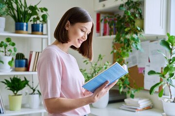 Happy smiling focused young woman reading book at home near window