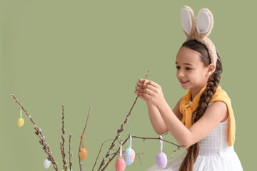 Cute little girl in bunny ears decorating willow branches with Easter eggs on green background