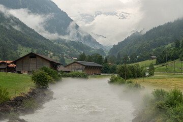 Fototapeta na wymiar View of an alpine river in the valley during a rainy summer day. Mountains on all sides, clouds hanging between the mountains. Spray is coming of the wild river. Mystic landscape in Switzerland.