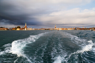 Venice panorama from sea and bright sea water trail behind small boat. San Giorgio Maggiore island, Saint Mark's Square, San Marco district view from water bus with dark blue stormy sky in background